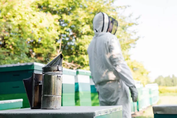 Selective focus of bee smoker near beehives and blurred beekeeper in protective suit — Stock Photo