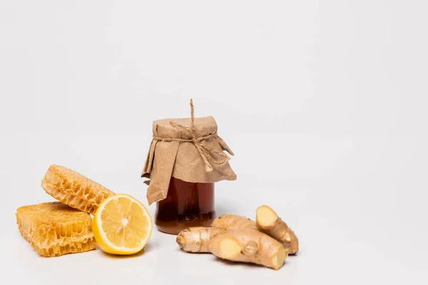 Pot de miel recouvert de papier artisanal près de citron frais, racine de gingembre et nid d'abeille sur blanc — Photo de stock