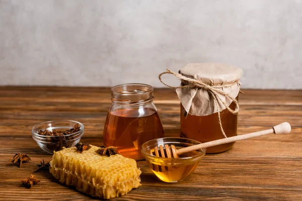 Jars with honey near honeycomb, anise seeds and dipper on wooden table and grey background — Stock Photo