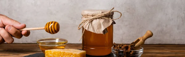 Vue recadrée de la main masculine avec trempette en bois près de nid d'abeille, pot et bols avec du miel et des graines d'anis sur fond gris, bannière — Photo de stock