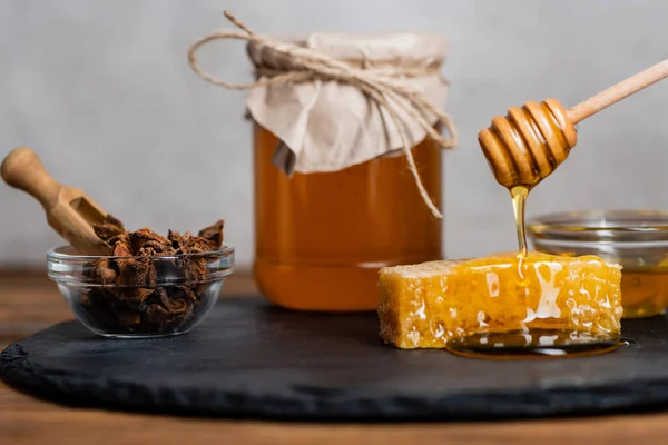 Nido d'ape, paletta di legno, ciotola con semi di anice e barattolo sfocato con miele fresco su lavagna e fondo grigio — Foto stock