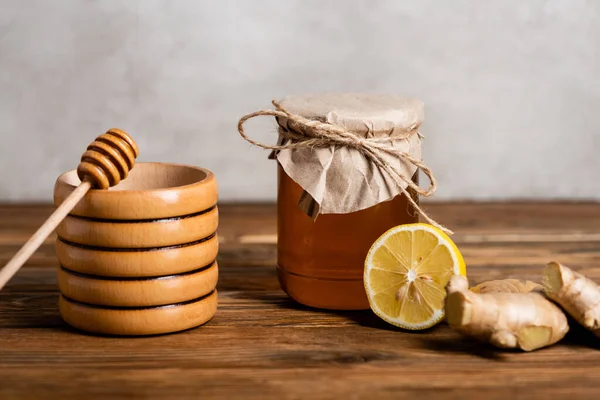 Récipient et trempette en bois près de la moitié du citron frais, racine de gingembre et pot avec du miel sur fond gris — Photo de stock