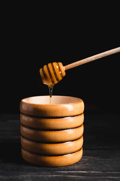 Liquid honey flowing from dipper into wooden container isolated on black — Stock Photo