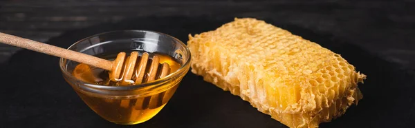 Bowl with honey and wooden dipper near honeycomb on black surface, banner — Stock Photo