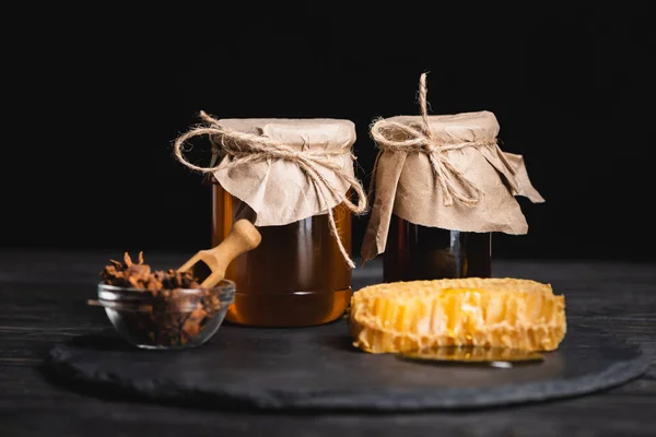 Honey jars covered with craft paper near bowl with anise seeds and honeycomb isolated on black — Stock Photo