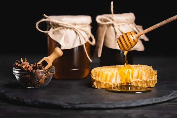 Mergulhador de madeira com mel líquido perto de favo de mel, tigela com sementes de anis e frascos borrados isolados em preto — Fotografia de Stock