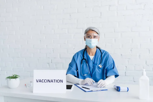 Middle aged asian doctor in goggles and medical mask near sign plate with vaccination lettering on desk — Stock Photo