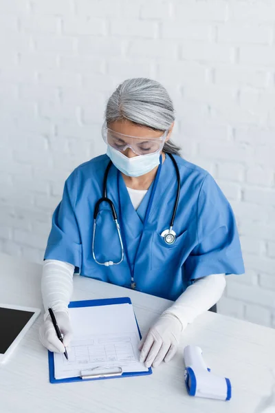 Immunologo asiatico in maschera medica, occhiali e guanti di lattice scrivendo su carta medica, concetto di vaccinazione — Foto stock