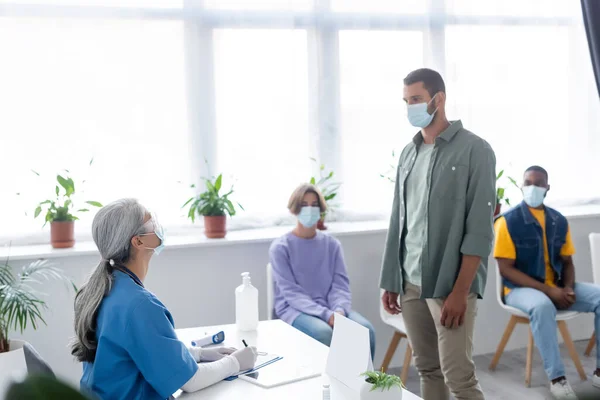 Middle aged nurse talking to man in medical mask near blurred multiethnic patients — Stock Photo