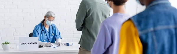 Madura asiática enfermera en médico máscara escritura en vacunación centro cerca borrosa multiétnica pacientes, bandera — Stock Photo