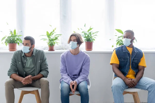 Молодые мультиэтнические мужчины в медицинских масках сидят в очереди в ожидании вакцинации — стоковое фото