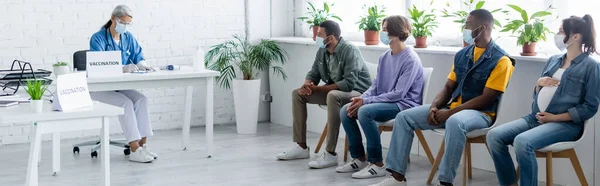 Reife Krankenschwester schreibt in Impfzentrum in der Nähe von Patienten, die in medizinischen Masken Schlange stehen, Transparent — Stockfoto