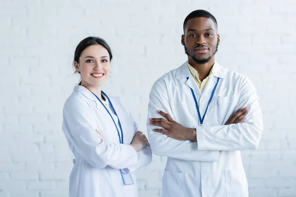 Positive multiethnische Ärzte in weißen Kitteln stehen mit verschränkten Armen da und lächeln in die Kamera — Stockfoto