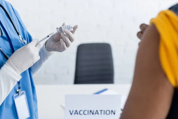 Частковий вигляд лікаря в латексних рукавичках, що тримає вакцину біля розмитого афроамериканця в центрі вакцинації — стокове фото