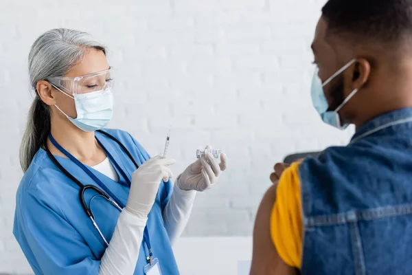 Asiatische Krankenschwester in medizinischer Maske hält Spritze und Gefäß mit Impfstoff in der Nähe verschwommener afrikanisch-amerikanischer Mann — Stockfoto