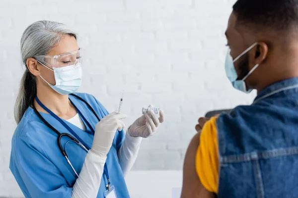 Азіатська медсестра в медичній масці тримає шприц і банку з вакциною поблизу розмитого афроамериканця — стокове фото