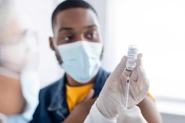 Медсестра в латексних рукавичках, що наповнюють шприц коронавірусною вакциною біля розмитого афроамериканця в медичній масці — стокове фото
