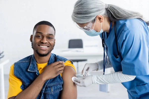 Asian doctor in medical mask vaccinating happy african american man in clinic — Stock Photo