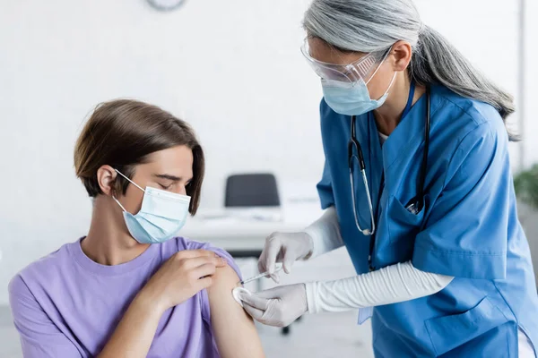 Mature asian doctor in medical mask and goggles giving vaccine injection to young patient — Stock Photo