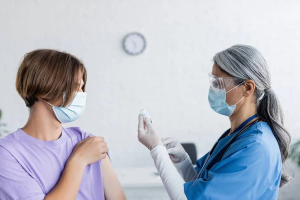 Young man in medical mask near mature asian nurse holding jar with vaccine — Stock Photo