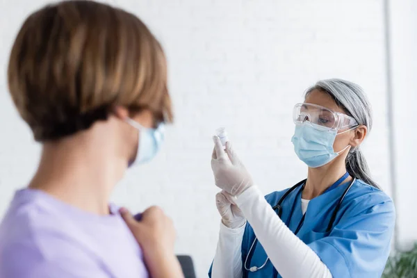 Verschwommener Mann in der Nähe reifer asiatischer Krankenschwester in medizinischer Maske und Latexhandschuhen, die ein Glas mit Impfstoff halten — Stockfoto