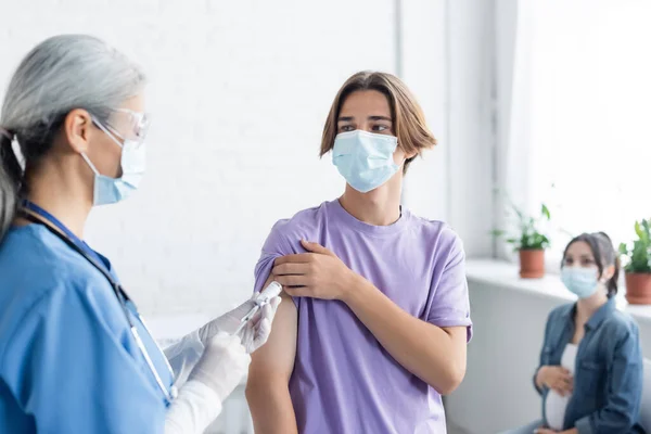 Médico en máscara médica y guantes de látex vacunando a un hombre joven cerca de la mujer embarazada borrosa - foto de stock