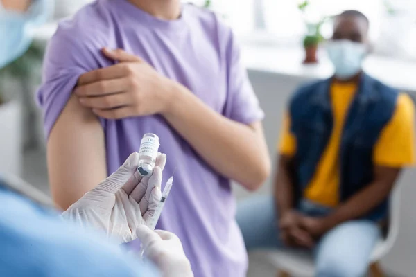Infirmière floue dans des gants en latex tenant le vaccin près du jeune patient et l'homme afro-américain — Photo de stock