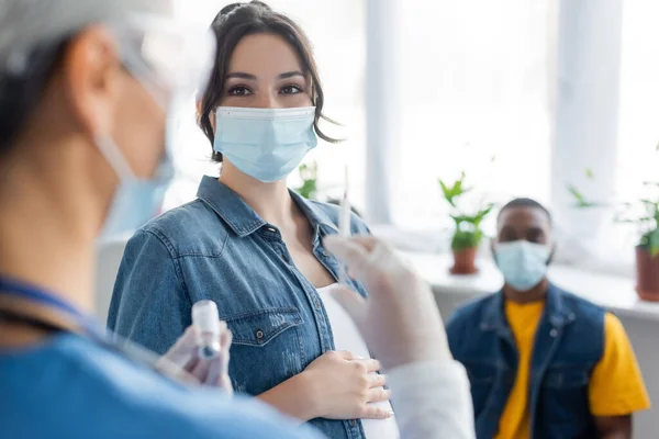 Enfermeira desfocada segurando seringa e frasco com vacina perto de mulher grávida e homem afro-americano em máscaras médicas — Fotografia de Stock