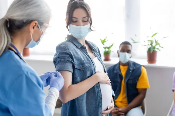 Зріла медсестра вакцинація вагітної жінки біля розмитого афроамериканця в медичній масці — стокове фото