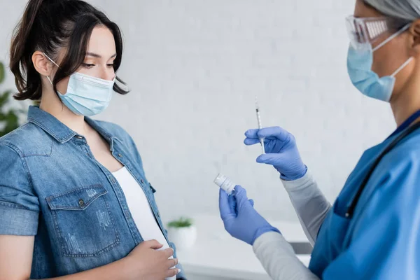 Médico asiático borroso en máscara médica y guantes de látex mostrando frasco con vacuna a la mujer embarazada - foto de stock