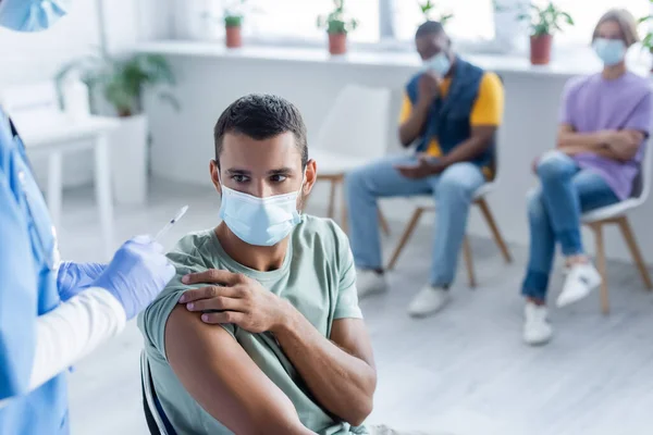 Врач держит шприц с вакциной рядом с человеком в медицинской маске и размытые мультикультурных пациентов — стоковое фото