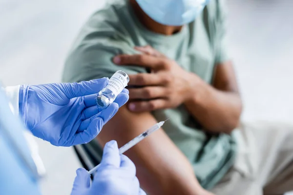 Vista ritagliata dell'uomo sfocato in maschera medica vicino al medico che tiene barattolo e siringa con vaccino — Foto stock