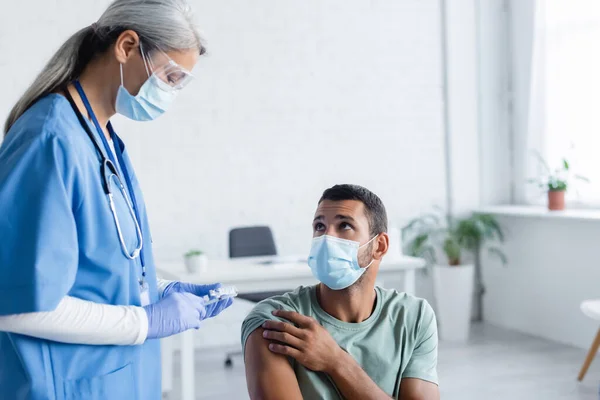 Mature asian nurse in medical mask, goggles and latex gloves holding vaccine near young man — Stock Photo