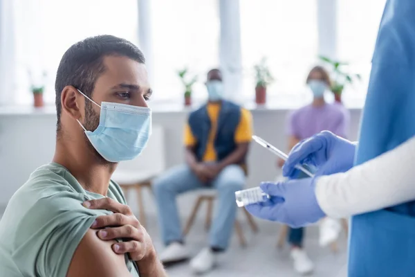 Enfermeira em luvas de látex segurando seringa e frasco com vacina perto do homem em máscara médica e pessoas multiétnicas borradas — Fotografia de Stock