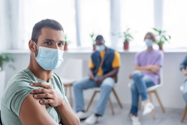Молодой человек в медицинской маске, смотрящий в камеру рядом с размытыми мультиэтническими людьми в центре вакцинации — стоковое фото