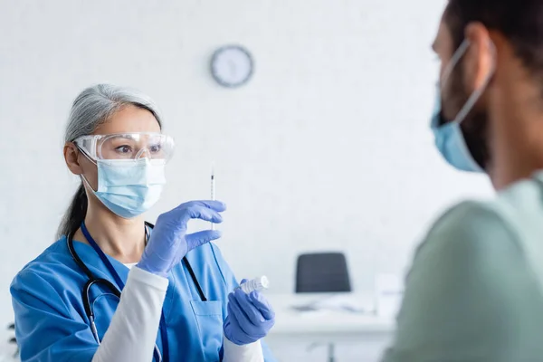 Médico asiático maduro en máscara médica y gafas preparar jeringa con vacuna cerca del hombre borroso - foto de stock