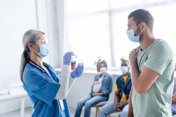 Mature asian nurse in medical mask holding vaccine near young man and blurred multiethnic patients — Stock Photo