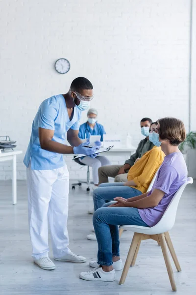 African american doctor in medical mask holding clipboard while talking to patients in vaccination center — Stock Photo