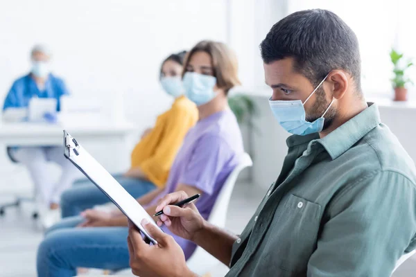 Joven en máscara médica escrito en portapapeles cerca de personas borrosas y enfermera, concepto de vacunación - foto de stock