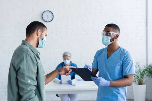 Homme dans le masque médical pointant vers presse-papiers dans les mains du médecin afro-américain dans le centre de vaccination — Photo de stock