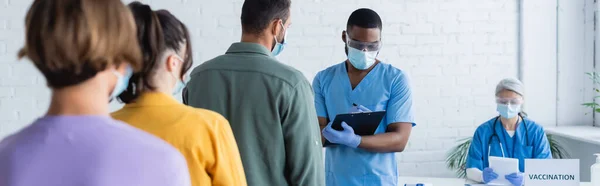 African american doctor in medical mask writing on clipboard near patients and blurred asian colleague, banner — Stock Photo