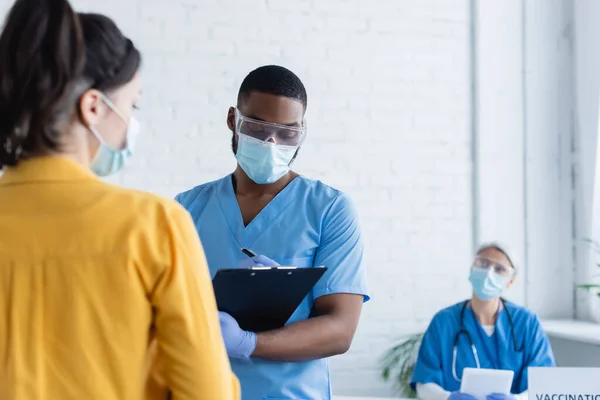 Afro-américain en masque médical écrit sur presse-papiers près de femme floue dans le centre de vaccination — Photo de stock