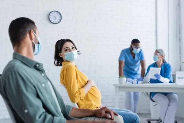 Mujer embarazada con máscara médica cerca del hombre y los médicos interracial borrosa en el centro de vacunación - foto de stock