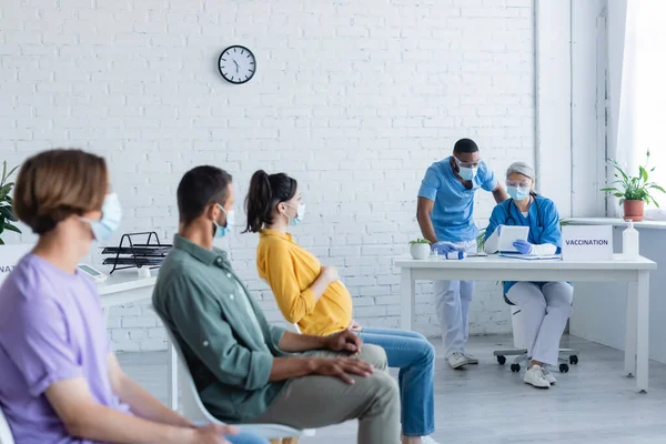 Mujeres embarazadas y hombres jóvenes con máscaras médicas sentados en el centro de vacunación cerca de médicos multiétnicos - foto de stock