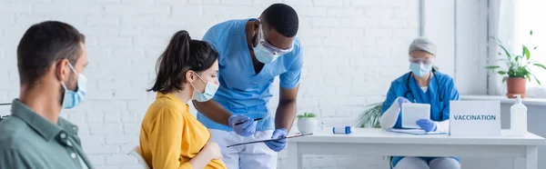 Médico afroamericano con portapapeles y pluma cerca de la mujer embarazada en el centro de vacunación, pancarta - foto de stock