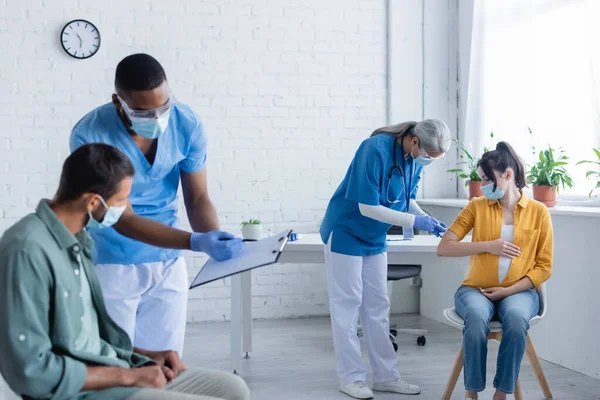 Multiethnic doctors in medical masks and uniform working with patients in vaccination center — Stock Photo