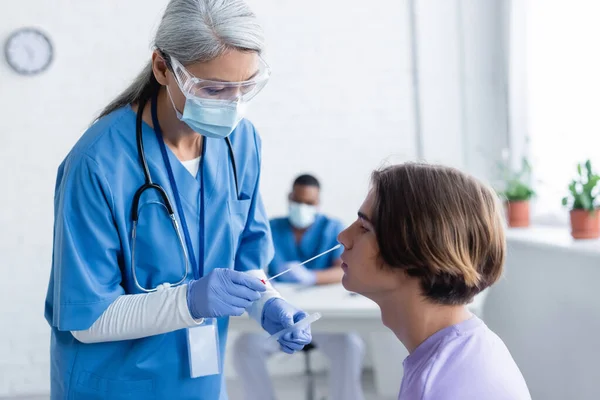 Mature asian doctor in medical mask and goggles making coronavirus test to young man — Stock Photo