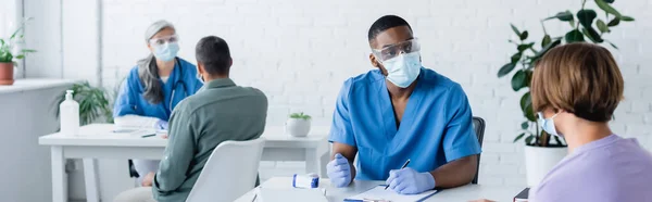 Médicos multiculturales en máscaras médicas hablando con los pacientes en el centro de vacunación, pancarta - foto de stock