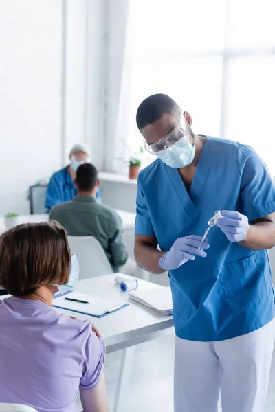 Médico afro-americano em máscara médica e luvas de látex preenchendo seringa com vacina — Fotografia de Stock