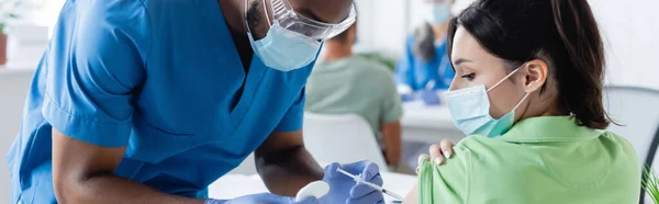 Африканский американский врач в медицинской маске вакцинирует молодую женщину в больнице, баннер — стоковое фото